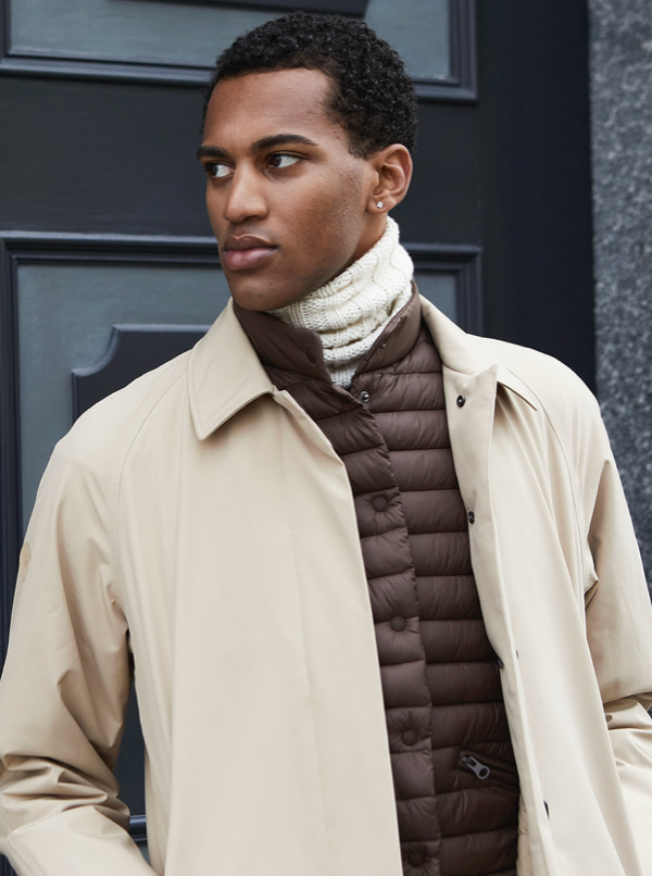 A man wearing a beige coat over a brown puffer jacket and a white ribbed scarf stands in front of a dark blue door, looking to his left.