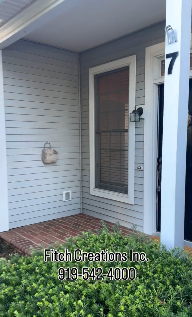 A light gray house exterior with brick steps, a black door labeled “7,” a window with blinds, a wall-mounted basket, and green shrubs in front. Text at the bottom reads: “Fitch Creations Inc. 919-542-4000.”.