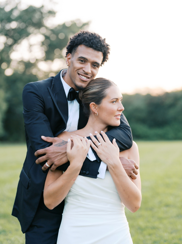 A man in a black suit embraces a woman in a white dress from behind. They stand outdoors on green grass with trees in the background, both smiling and appearing happy.