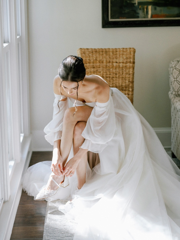 A bride in an off-the-shoulder white wedding gown sits on a wicker chair by a window, bending forward to fasten her shoe, with soft natural light illuminating the scene.