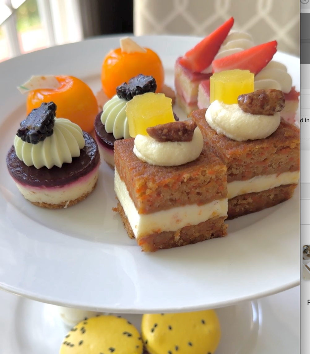 An assortment of elegant mini desserts on a white tiered stand, including carrot cake squares, fruit-topped pastries, and other colorful sweets arranged neatly.