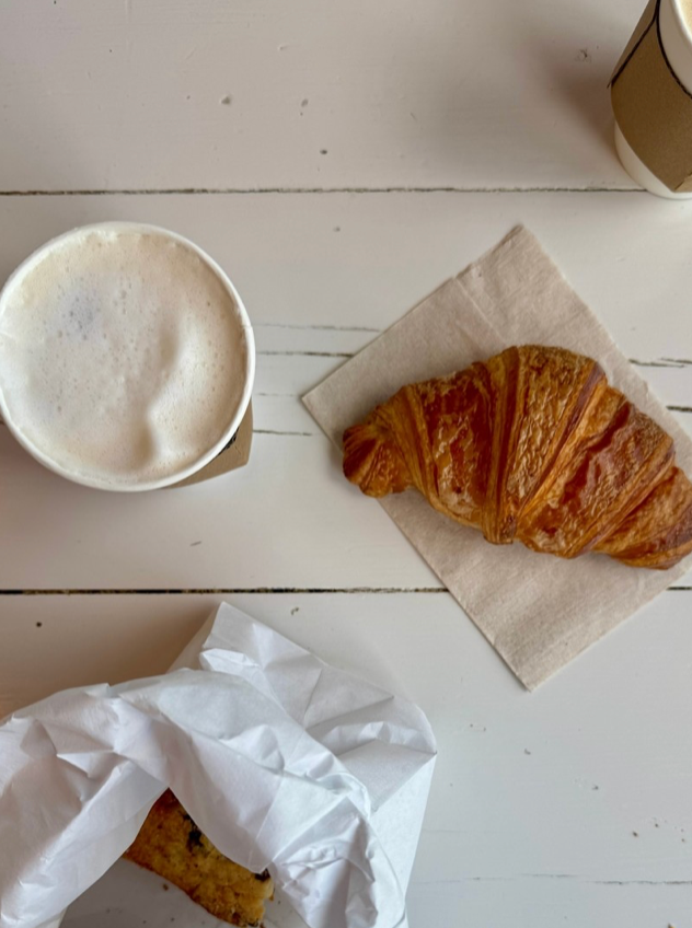 A cup of frothy coffee, a croissant on a napkin, and a partially wrapped pastry on a white wooden table, with another takeaway coffee cup partially visible in the corner.