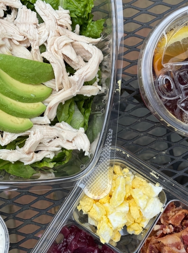 A close-up of a meal on a metal table, showing a salad with sliced avocado and shredded chicken, a divided tray with scrambled eggs, bacon, and beets, and a plastic cup of iced tea with lemon.