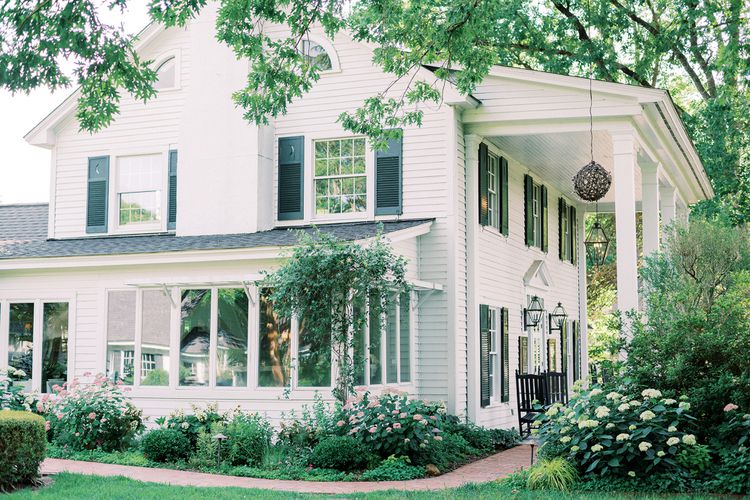 A white two-story house with black shutters and a covered porch, surrounded by lush green trees, bushes, and blooming flowers along a brick pathway.