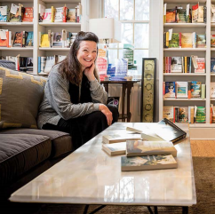 A woman sits smiling on a dark sofa in a cozy room filled with bookshelves. A marble coffee table in front of her holds several open books. Sunlight streams through a window behind her.