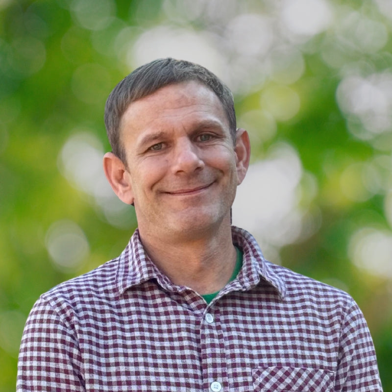 A man wearing a red and white checkered shirt smiles gently at the camera, standing outdoors with a blurred background of green foliage and dappled sunlight.