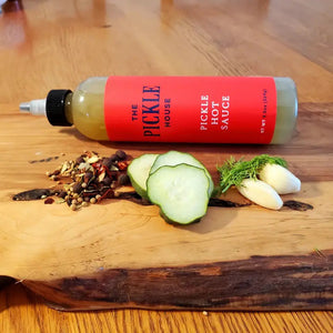 The product labeled "PICKLE HOUSE - PICKLE HOT SAUCE" from THE PICKLE HOUSE is displayed against a plain white background. The bottle, adorned with a red label featuring black text and capped with a white nozzle, contains 9.8oz (280g) of greenish-hued sauce. Its distinctive flavor arises from a secret spice blend mixed with tangy pickle brine.