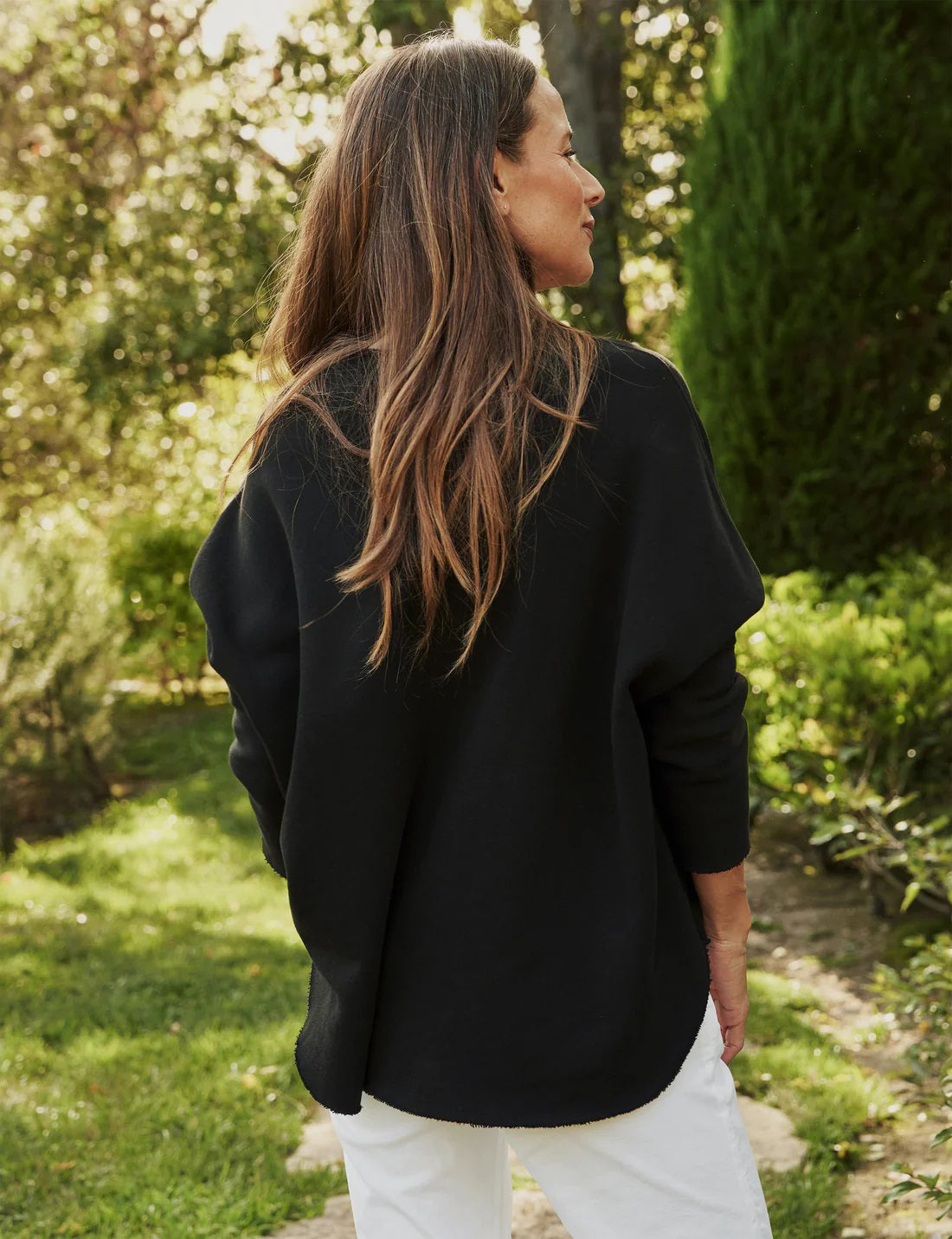 A person with long, wavy blonde hair is standing outdoors near a large tree, wearing the FRANK &amp; EILEEN ANNA LONG-SLEEVE CAPELET in navy over a white shirt and blue pants. The background showcases green foliage and a stone path. The person is smiling and gazing to the side.