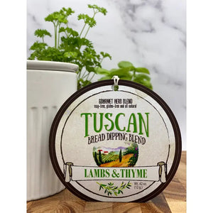 A circular label with the text "LAMBS & THYME - TUSCAN BREAD DIPPING BLEND" hangs in front of a white container holding a green leafy herb plant. The label's background features an illustration of a picturesque Tuscan countryside, perfectly setting the scene for your gourmet appetizer.