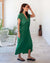 A woman stands in a well-lit hallway, wearing the vibrant MERSEA - IBIZA KAFTAN DRESS, accessorized with a necklace and black sandals. The lightweight green dress is complemented by a white chair and greenery in the background, creating a resort-worthy, modern atmosphere.