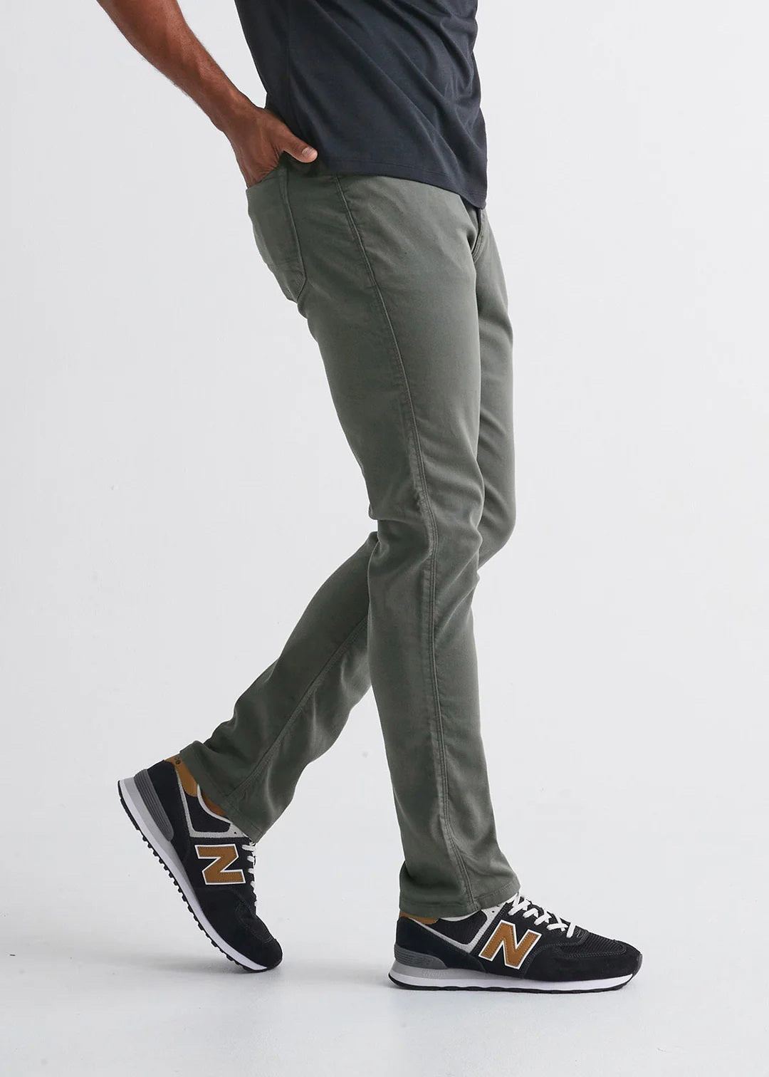 A person stands against a plain white background wearing DUER No Sweat Relaxed Taper Pants in Gull (olive green), a black T-shirt, and black New Balance sneakers with white soles and gold accents.