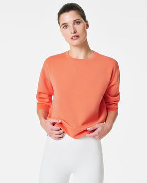 A woman stands confidently against a plain white background, wearing the SPANX Aireessentials Crew Neck Pullover and matching sweatpants made from luxurious comfort spacer fabric. She has her hair pulled back and her left hand is raised slightly. She smiles gently with her lips closed.