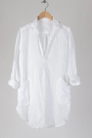 A long-sleeved, dark blue CP SHADES - TETON TUNIC from the brand CP SHADES hangs on a white hanger against a light background. The 100% linen tunic features a collar, V-neckline opening, and large front pockets. A white tag with text is attached to the neckline.