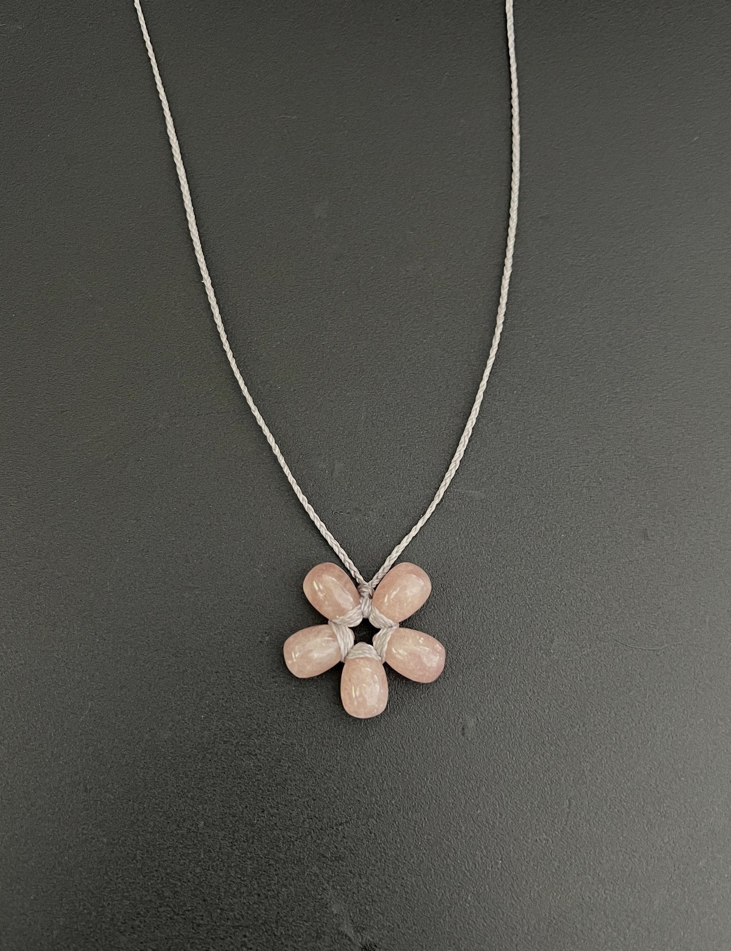 The ANN LIGHTFOOT Petite Sunstone Flower Necklace features a flower-shaped pendant with five pink stone petals and a central white stone on a dark setting. It has a delicate silver chain and adjustable nylon ties for the perfect fit.