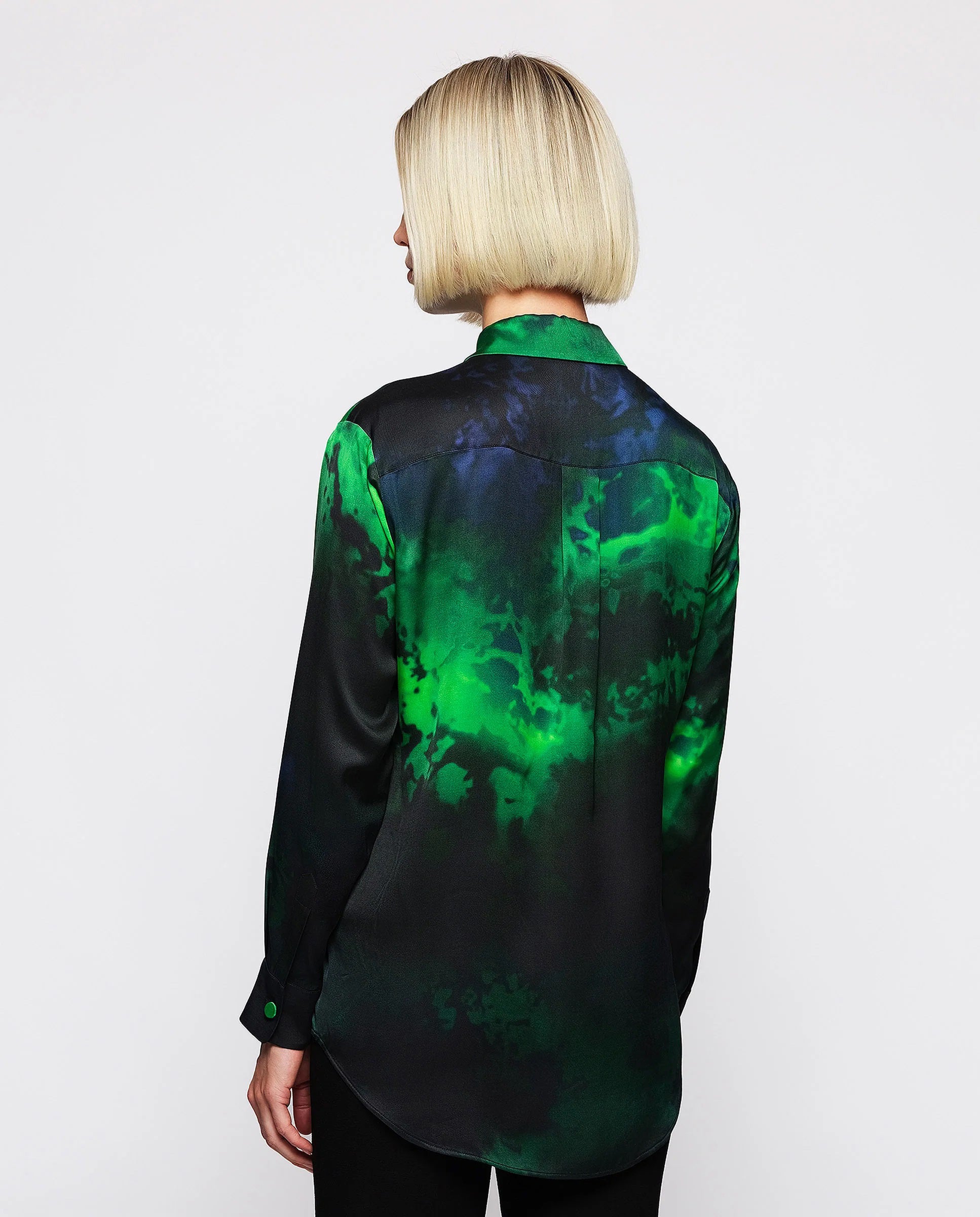 A person with a blonde bob haircut is wearing the MIRTO 1956 - GREEN AND BLACK PRINT FLUID BLOUSE, a long-sleeve, button-up featuring an exclusive green and black abstract pattern. The brand name MIRTO 1956 is highlighted in the design. The person stands against a plain white background, looking slightly to the side with a neutral expression.