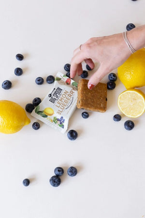 A white packet of Big Spoon Roasters Lemon Blueberry Cashew Snack Bar features colorful illustrations of lemons, blueberries, and cashews on the front. The packaging indicates that it is vegan, dairy-free, gluten-free, soy-free, and palm oil-free, with a net weight of 60g (2.12 oz).