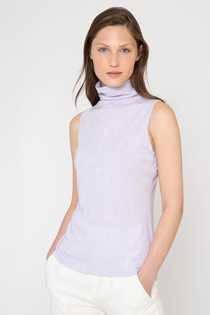 A woman with long brown hair is wearing the KINROSS Sleeveless Seamed Funnel top in light purple, paired with white pants. The luxurious outfit, crafted from a blend of silk and cashmere, exudes elegance as she stands with her hands in her pockets against a plain light background.