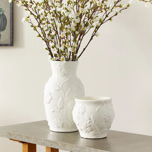 A decorative scene featuring the BLOSSOM CACHEPOT by NAPA HOME AND GARDEN, a tall white ceramic vase adorned with floral stem designs, filled with blooming white flowers on long stems. Next to it is a shorter, similarly patterned white ceramic jug on a wooden table with a gray surface, creating an elegant look.