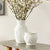 A decorative scene featuring the BLOSSOM CACHEPOT by NAPA HOME AND GARDEN, a tall white ceramic vase adorned with floral stem designs, filled with blooming white flowers on long stems. Next to it is a shorter, similarly patterned white ceramic jug on a wooden table with a gray surface, creating an elegant look.