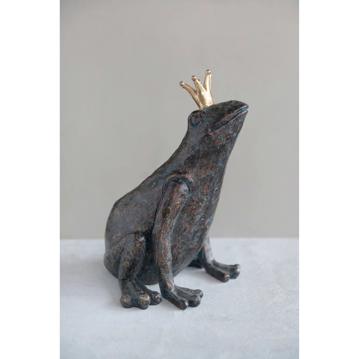 The CREATIVE COOP RESIN FROG WITH GOLD CROWN is a decorative sculpture in bronze patina finish, sitting upright with a gold crown, textured surface, and slightly weathered appearance as it looks upward.