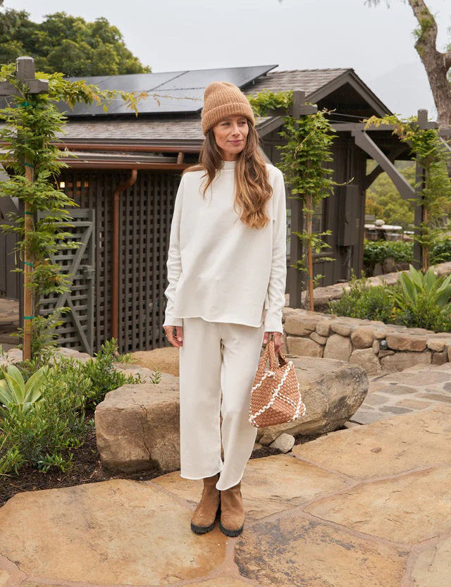 A woman with long brown hair stands smiling near a black grand piano in a modern, well-lit room. She wears the FRANK AND EILEEN - EFFIE LONG SLEEVE FUNNEL NECK CAPELET from FRANK &amp; EILEEN paired with blue jeans. A large window and a lamp are in the background, enhancing the cozy winter warmth of her stylish ensemble.
