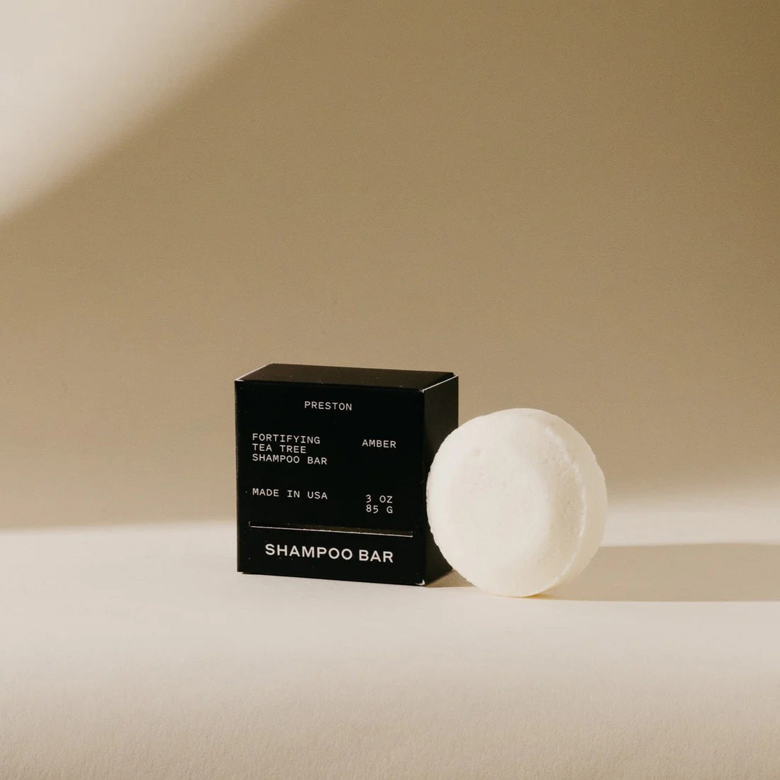 A small, black rectangular box of sulfate-free fortifying tea tree shampoo bar stands against a neutral beige background. The packaging reads &quot;PRESTON - SHAMPOO BAR, Fortifying Tea Tree Leather Shampoo Bar, Made in USA, 3 oz, 85.6 g,&quot; highlighting its plastic-free and plant-based cleansers.