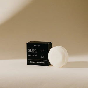 A small, black rectangular box of sulfate-free fortifying tea tree shampoo bar stands against a neutral beige background. The packaging reads "PRESTON - SHAMPOO BAR, Fortifying Tea Tree Leather Shampoo Bar, Made in USA, 3 oz, 85.6 g," highlighting its plastic-free and plant-based cleansers.