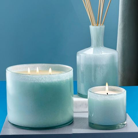 A serene setup with a LAFCO Marine Bathroom 3 Wick Candle and a smaller soy candle, both lit, beside a blue reed diffuser. The soft blue background evokes a calming sea air atmosphere.