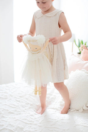 A young girl with light hair tied in a bow sits on a bed with white linens, smiling and holding up an Alimrose Ava Angel Doll Ivory Gold. She wears a light-colored dress, and behind her are pink and rust-colored pillows against a gray headboard.