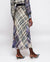 A person is modeling an uncomplicated chic outfit that includes the MIRTO 1956 - BLUE PLAID MIDI SKIRT paired with a long-sleeved, high-necked blouse, pointed heels, and holding a small, dark handbag. The skirt features a cinched waist and long design, and the person has short hair.