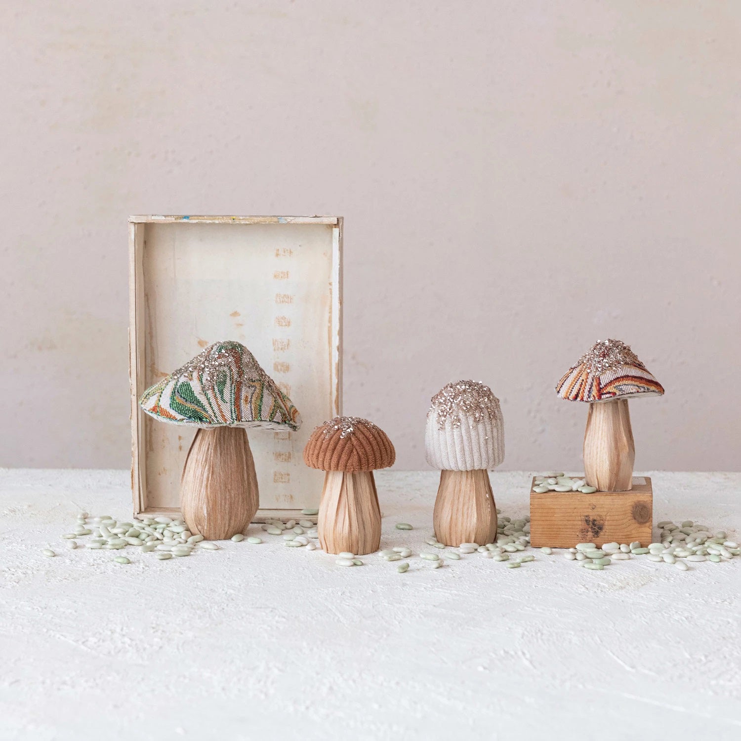 Four delightful mushrooms with fabric tops are elegantly arranged on a wooden base, each varying in height and design. Their earthy tones contrast beautifully with subtle multi-color hues, embodying the Creative Co-Op charm. A rectangular wooden box stands open in the background, while small seeds are artfully scattered around the display.