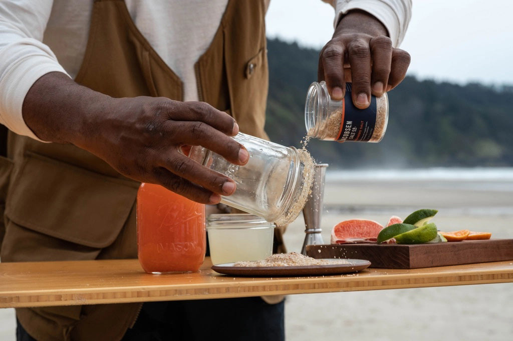 A person is preparing a cocktail at an outdoor setup on a beach. They are pouring Jacobsen Salt Co.'s Jacobsen Salt - Infused Habanero Salt onto a plate from a jar. The setup includes jars of liquid, a small tray with fruit slices, and a scenic backdrop of trees and sand.