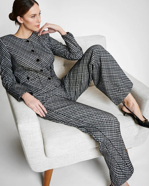 A person with long hair sits casually on a light-colored chair wearing the SPANX PONTE JACQUARD JACKET, which is a black and white checkered suit adorned with gold-finished buttons. They are gazing to the side, with one arm resting on the chair's armrest and the other in their lap.