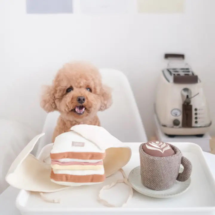 A small brown poodle sits at a table with a stuffed toy cake and a stuffed toy cup of latte art. The background features a white wall and a coffee maker. The poodle, eager for its LAMBWOLF COLLECTIVE - HAM AND CHEESE DOG TOY burrow challenge, looks directly at the camera with a happy expression.