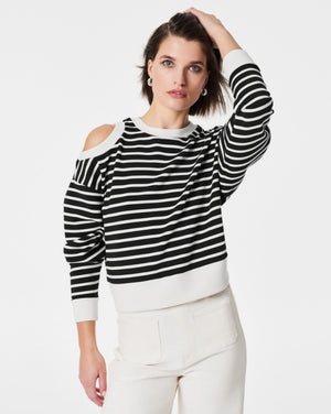 A person with short hair poses against a white background, wearing the SPANX - AIRESSENTIALS COLD SHOULDER TOP in black and white stripes crafted from luxuriously soft material, paired with light-colored pants. Their left hand is placed on their head while their right arm rests at their side.