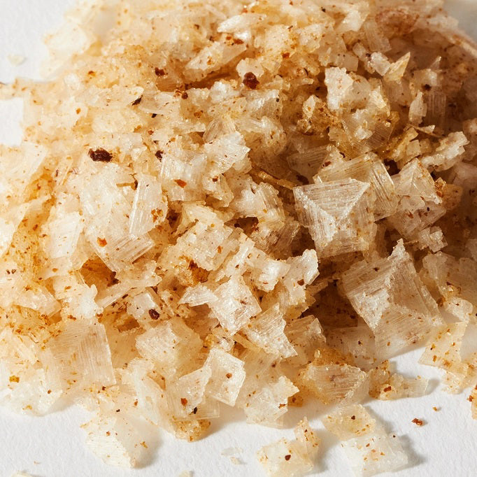 Close-up image of a small pile of JACOBSEN SALT - INFUSED WHITE TRUFFLE SALT on a white surface. The salt flakes, bearing the signature touch of JACOBSEN SALT CO., are irregularly shaped and exhibit a light golden-brown color, indicating the presence of white truffle infusions. The texture is coarse and flaky, making it perfect for garnishing seared beef tenderloin.