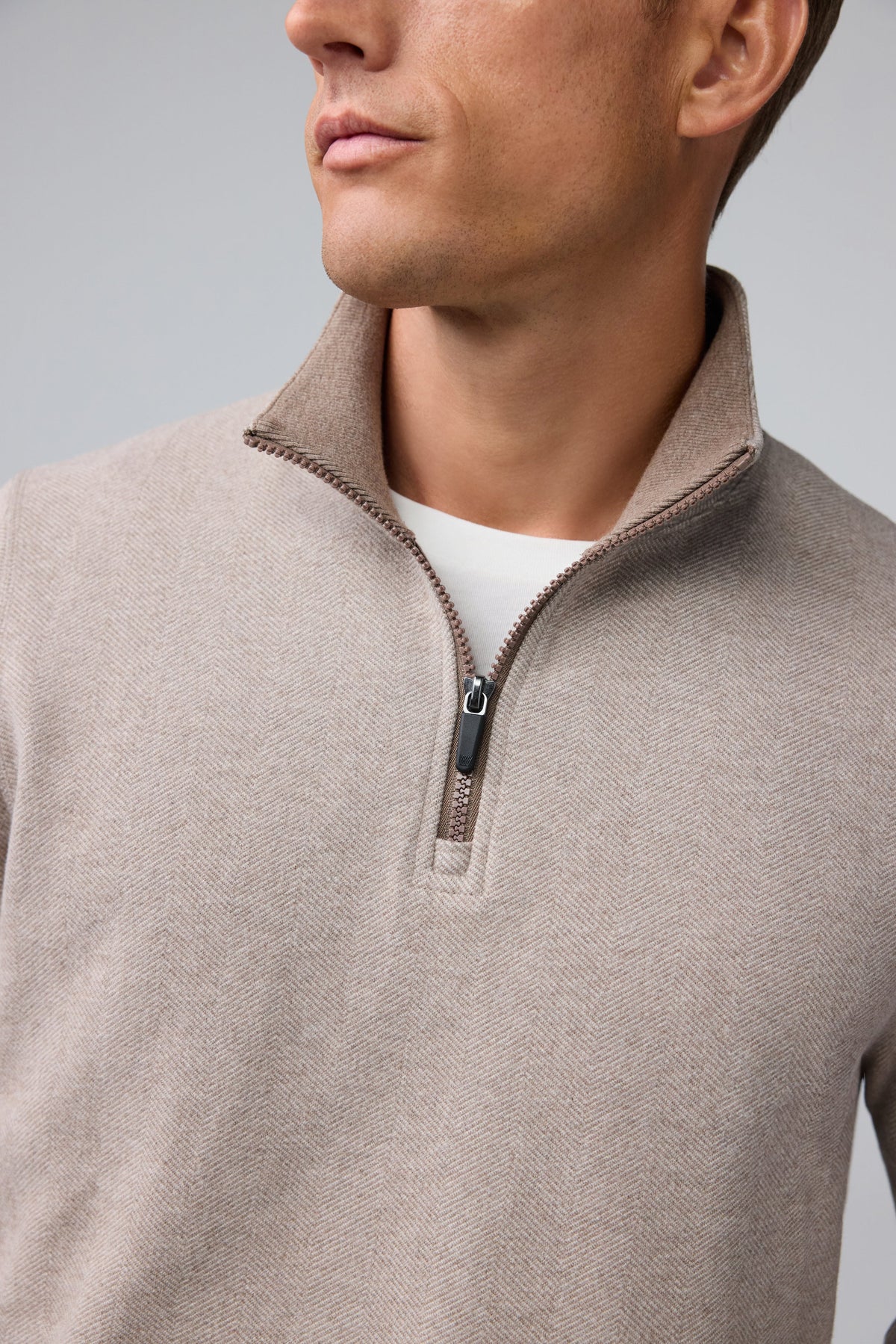 A man with short light brown hair stands in profile, wearing the MACK WELDON Ace 1/2 Zip Sweater in Herringbone and dark pants, one hand in his pocket, against a plain light gray background.