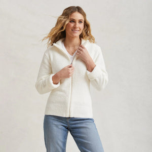 A person with long wavy hair is wearing a white, textured Kashwére Full Zip Sport Jacket and blue jeans. The individual is facing slightly to the viewer's left and smiling gently, with one hand adjusting the jacket's collar. The background is plain gray.