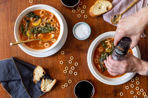 Two bowls of tomato-based soup with pasta and greens sit on a wooden table. A person seasons one bowl with pepper from a Jacobsen Salt Co. - Grinder Tellicherry Peppercorns. Slices of baguette, a cloth napkin, and glasses of red wine are placed around the bowls.