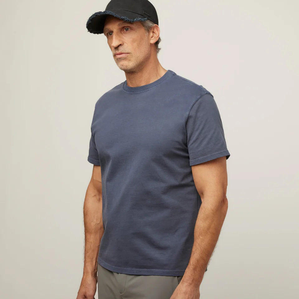 A man in a TILLEY MID WEIGHT SUNWASHED T-SHIRT IN MARINE NAVY, gray pants, and a black cap stands against a plain light background, gazing slightly to the side with a neutral expression.