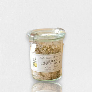 A small glass jar contains BELLA CUCINA - CITRUS & FENNEL SAVORY SALT, adorned with a "Citrus & Fennel" label featuring a lemon illustration. Enhanced by oven-dried citrus peel and fragrant fennel seeds, the jar is topped with a metal clasp and clear lid.