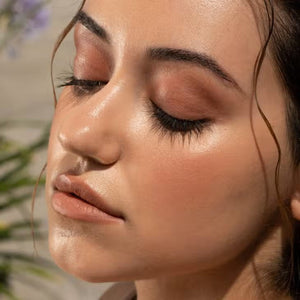 A close-up of a person with curly dark hair, a serious expression, and warm makeup featuring the universally flattering, weightless JASON WU - HOT FLUFF LIP/EYE/CHEEK by JASON WU BEAUTY on their face. They are looking directly at the camera with a neutral background.