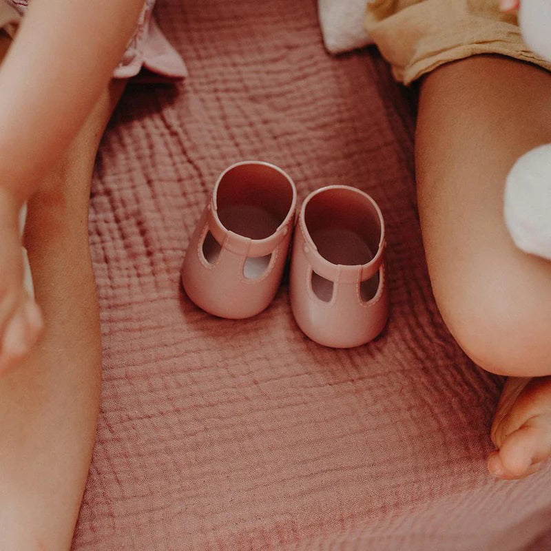 A close-up of a baby wearing OLLI ELLA USA DINKUM DOLL SHOES in Mallow Pink and white socks. The baby is seated on a soft, pale pink textured blanket, dressed in light brown clothing. Only the lower half of the baby&#39;s body is visible.
