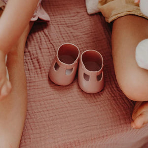 A close-up of a baby wearing OLLI ELLA USA DINKUM DOLL SHOES in Mallow Pink and white socks. The baby is seated on a soft, pale pink textured blanket, dressed in light brown clothing. Only the lower half of the baby's body is visible.