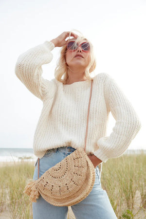 A person with blonde hair, wearing a white knit sweater and light blue jeans, stands outdoors on a grass-covered area near the beach. They are carrying the MAR Y SOL - LILA CROSSBODY bag from MAR Y SOL, which features a removable tassel. One hand is adjusting their sunglasses while the other rests on their hip.