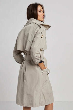 A woman with long brown hair poses in the ADROITE ATELIER - RAPHAEL TRENCH COAT by ADROIT ATELIER. This light beige trench coat highlights rolled-up sleeve details, a belted waist, and a flap on the back. She stands against a plain gray background, glancing over her shoulder.