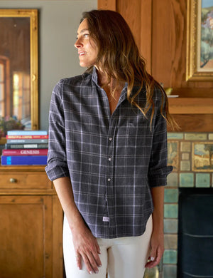 A woman in a bust-flattering FRANK & EILEEN Eileen Relaxed Button Up Shirt in Charcoal and Blue Plaid Italian Flannel and white pants stands indoors, looking left. She has long brown hair, and the room features a wooden cabinet with books and tiled fireplace, complementing her relaxed fit attire.