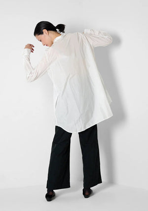 A person with curly hair wears the KATHARINA HOVMAN oversized taffeta blouse, featuring mother-of-pearl buttons, along with black pants. They stand in a minimalist setting with hands in pockets, gazing at the camera.