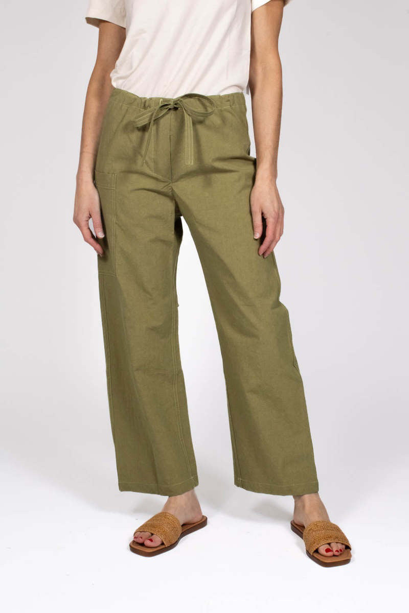 Against a plain background, a person wears THE LITTLE PROJECT's AMBER PANT in olive-green, made from Japanese cotton with a drawstring waist. They're styled with a white short-sleeve top and brown woven flat sandals. These versatile pants are already becoming a best seller.