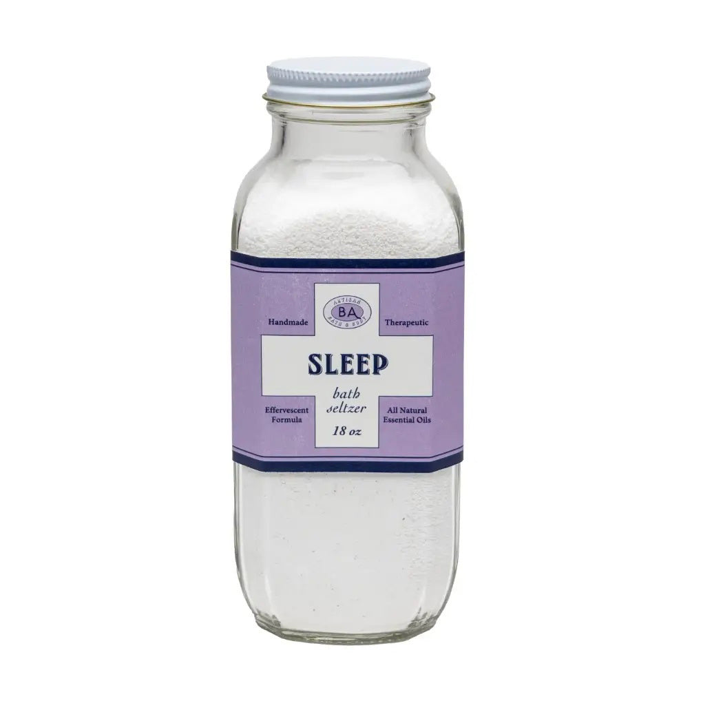 A clear glass jar labeled &quot;BAUDELAIRE - SLEEP BATH SELTZER&quot; features a purple label with ingredients, a barcode, and essential oils. Filled with granular white bath salts, its decorative border suggests instructions for a calming soak.